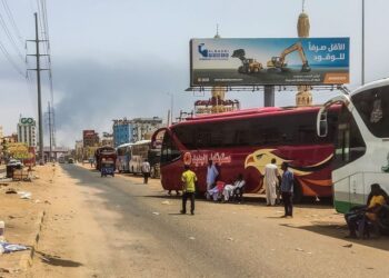 People wait next to passenger buses as smoke billows in an area in Khartoum where fighting continues between Sudan’s army and the paramilitary forces on April 28, 2023. – Fighting raged in Sudan on April 28, despite rival forces agreeing to extend a truce aimed to stem nearly two weeks of warfare that has killed hundreds and caused widespread destruction. (Photo by – / AFP)